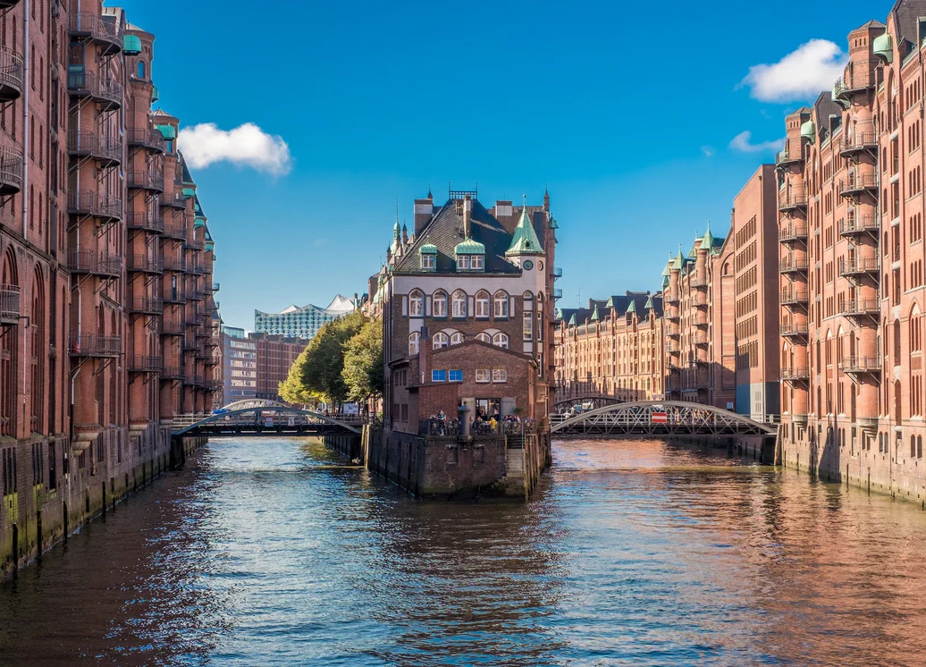 Historic brick buildings lining a canal with a small central building between two bridges, …